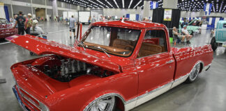 Front three-quarter high angle of a slammed candy red Chevrolet C10 pickup with an open hood showcasing an eight-stack velocity stack engine and chrome wheels inside the expo center.