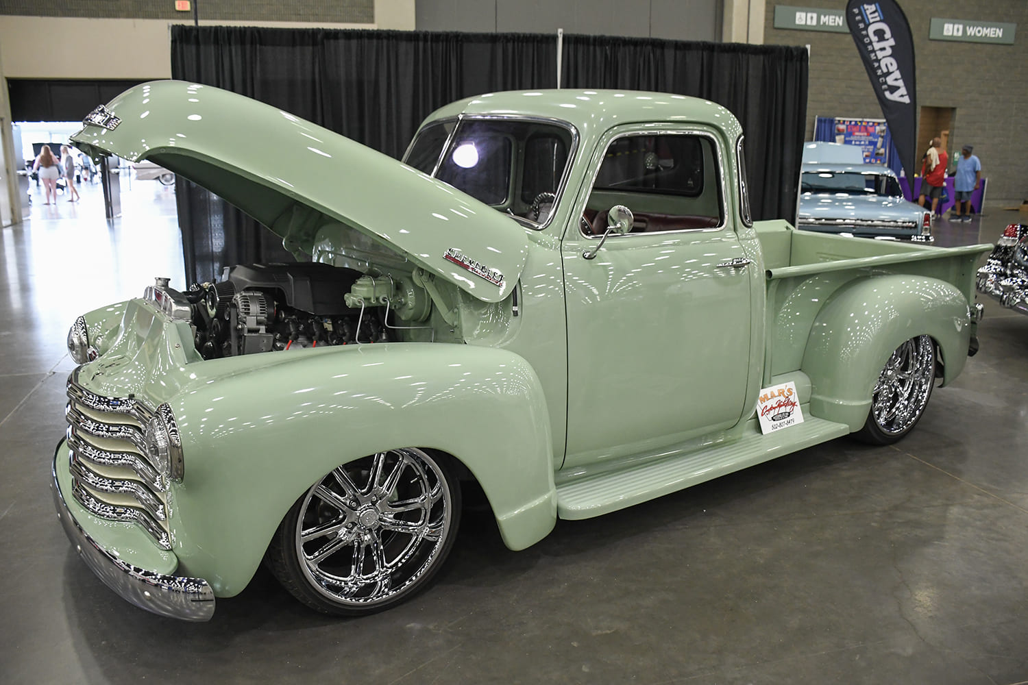 Front three-quarter angle of a custom pastel green vintage Chevrolet 3100 pickup truck with its butterfly hood propped open revealing a modern engine and polished chrome wheels.