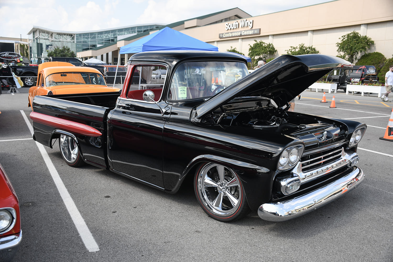 Front three-quarter view of a lowered black Chevrolet Apache pickup with an open hood, red side spear accents, and chrome five-spoke wheels in a show parking lot.