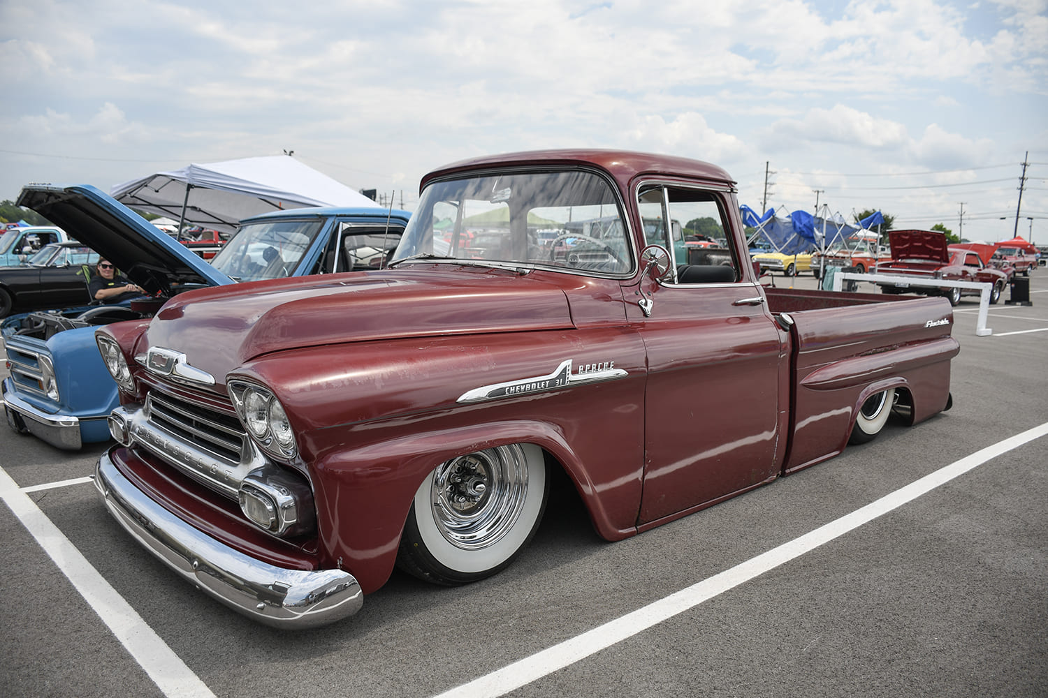 Low front three-quarter view of a metallic burgundy Chevrolet Apache fleetside pickup truck laid out low on chrome wheels with wide whitewall tires.
