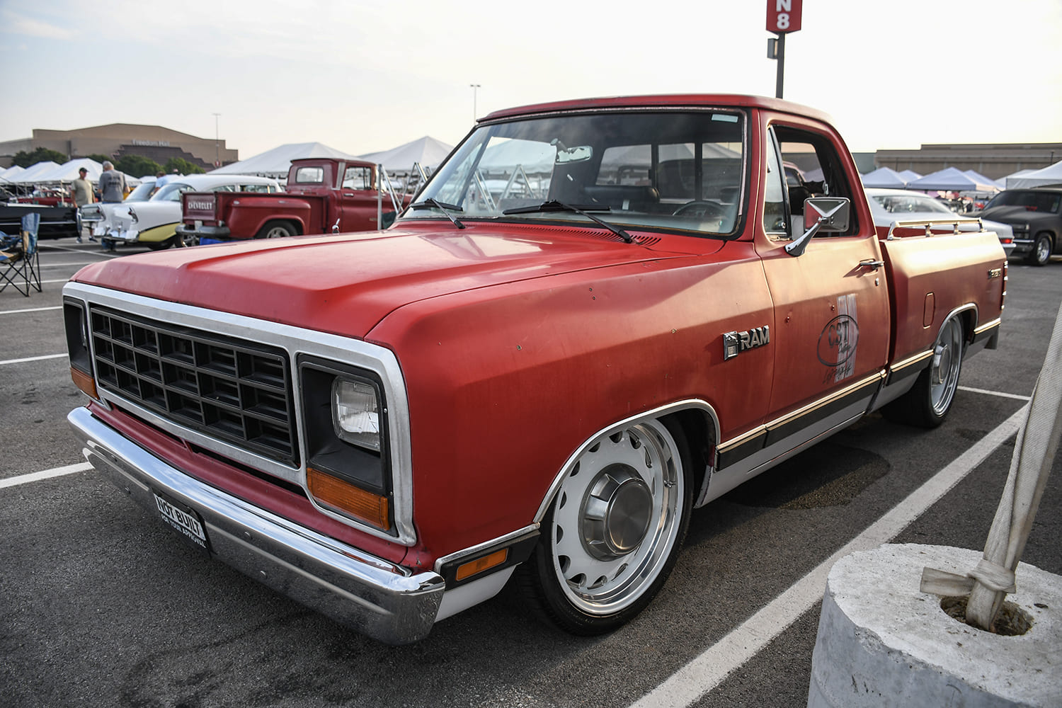 Front three-quarter perspective of a lowered red vintage Dodge Ram D-series pickup truck with painted solid wheels parked in an outdoor exhibition lot.