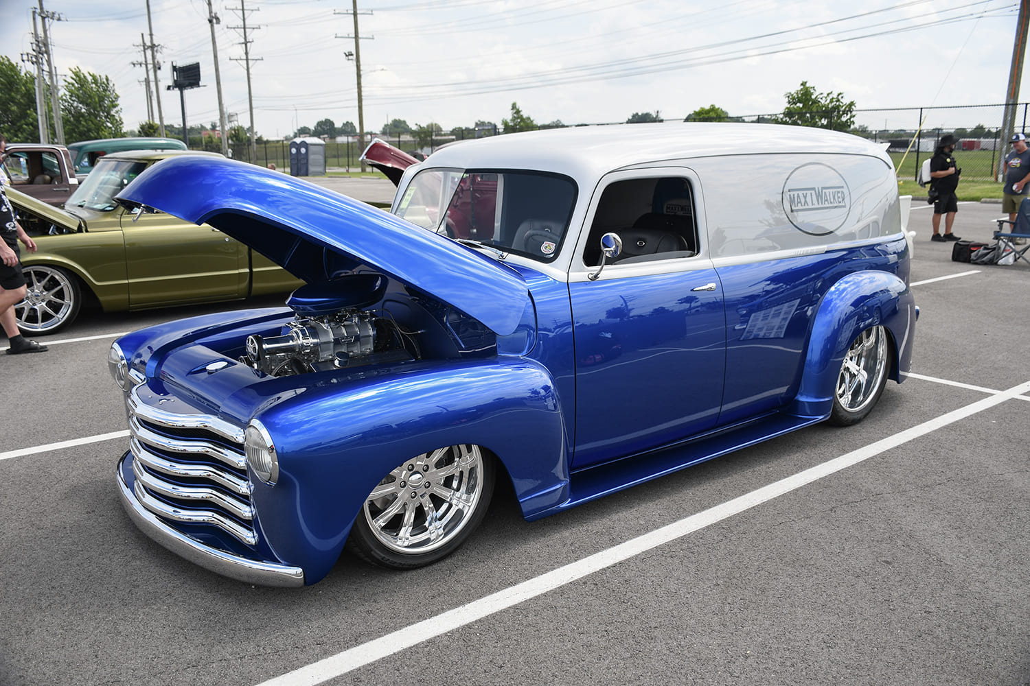 Front three-quarter view of a metallic royal blue and silver custom Chevrolet panel delivery truck with an open butterfly hood showcasing an engine with velocity stacks.