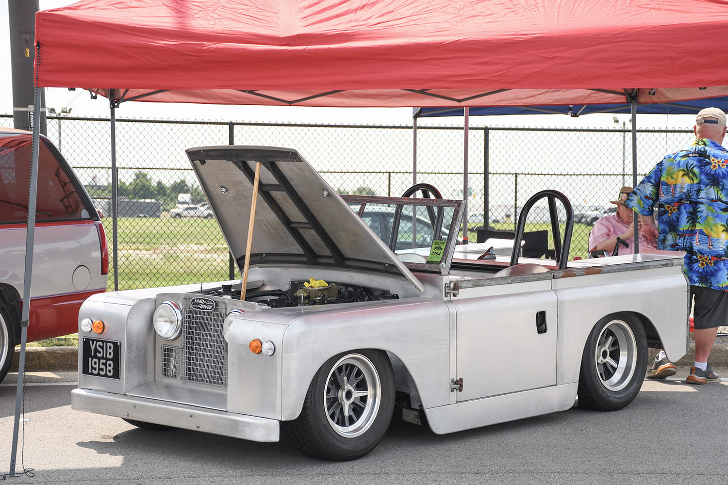 Front three-quarter view of a Cobra-inspired vintage Land Rover pickup in raw aluminum finish with an open hood under a red event tent canopy.