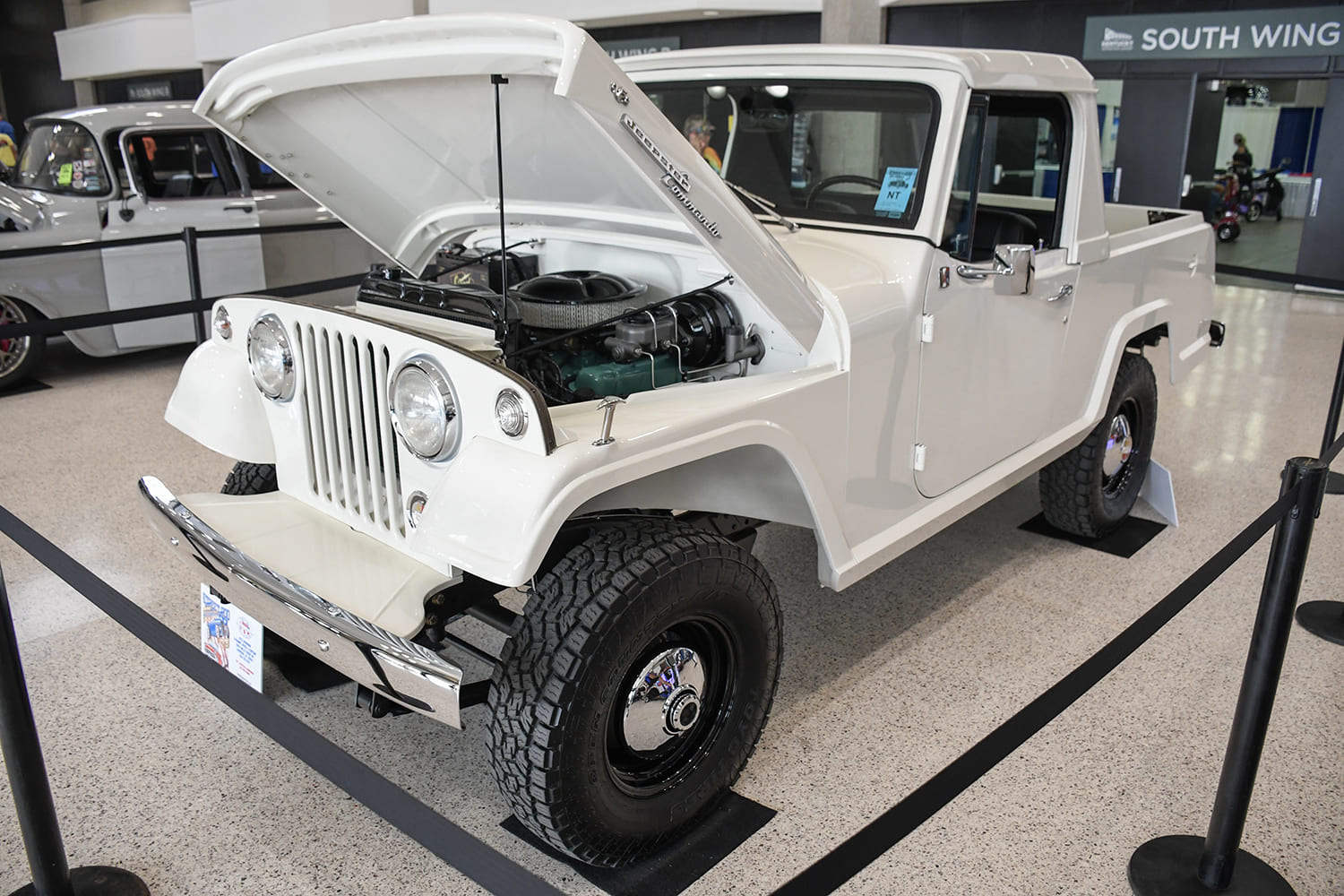 High front three-quarter view of a fully restored white Kaiser Jeepster Commando pickup with an open hood and all-terrain tires displayed inside the South Wing convention center.