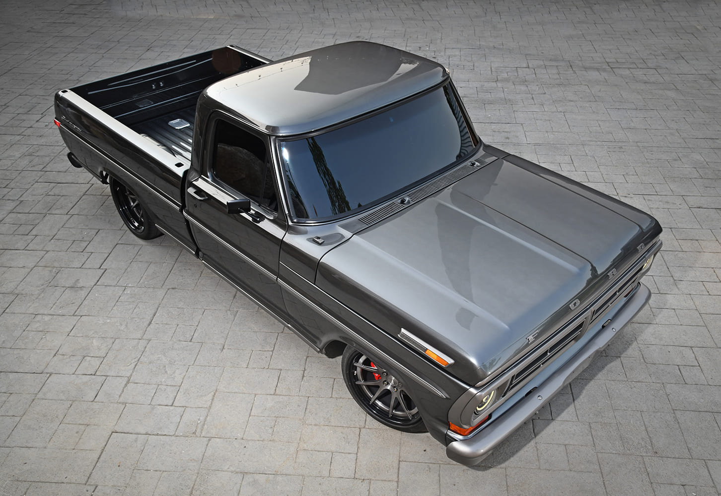 High aerial three-quarter shot of a gloss metallic charcoal gray 1972 Ford F-100 pickup truck with dark tinted windows and black multi-spoke wheels on stone pavers.