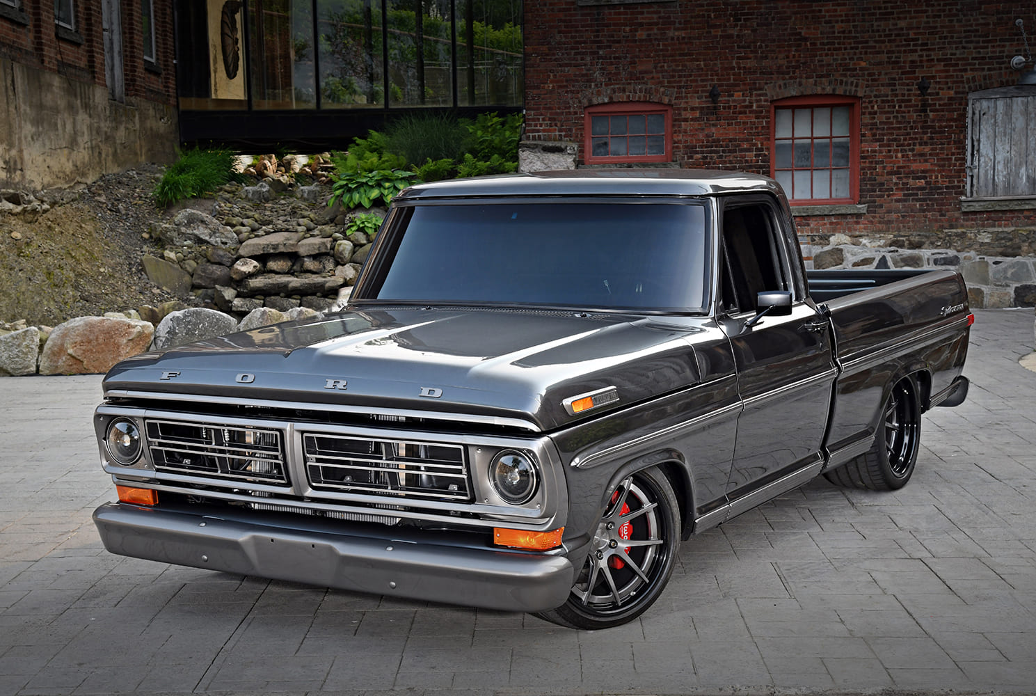 Front three-quarter view of a lowered 1972 Ford F-100 Sport Custom pickup in dark gray metallic paint parked in front of a rustic brick building.