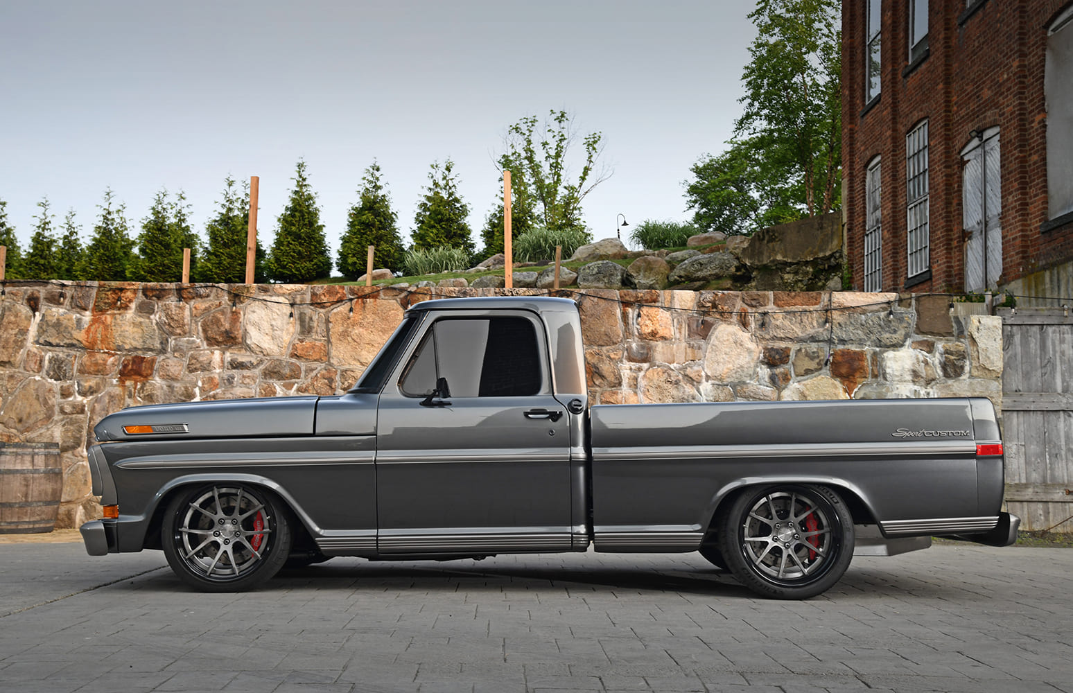 Complete side profile of a slammed dark metallic gray 1972 Ford F-100 Sport Custom showing a lowered suspension and red brake calipers against a stone wall.