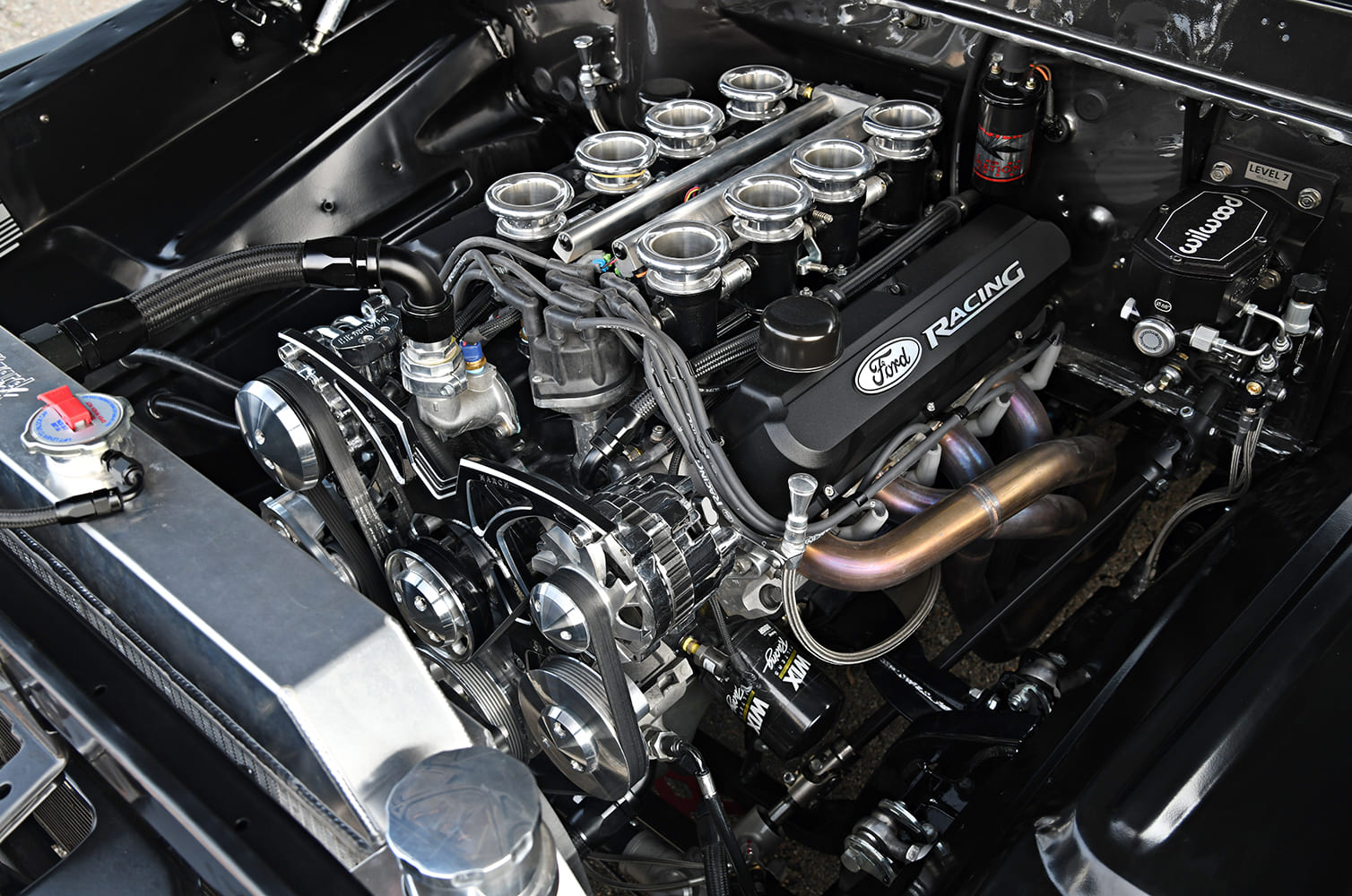 High front-angle view of a customized Ford Racing V8 with ITB injection, tubular headers, serpentine belt setup, and Wilwood brake booster.