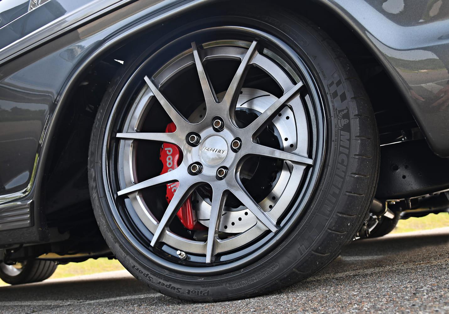 Close-up of a dark gray split-spoke Forgeline wheel wrapped in Michelin Pilot Super Sport tires with drilled and slotted Wilwood disc brakes and red calipers.