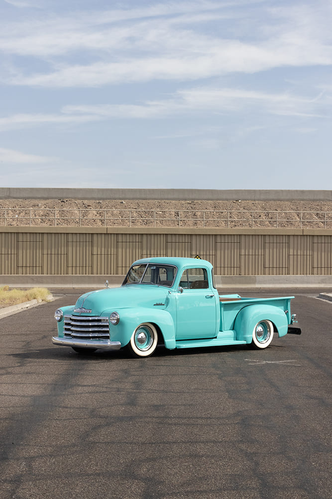 Front driver-side three-quarter profile of a lowered turquoise 1953 Chevy 3100 pickup truck parked on an open asphalt road under a blue sky.