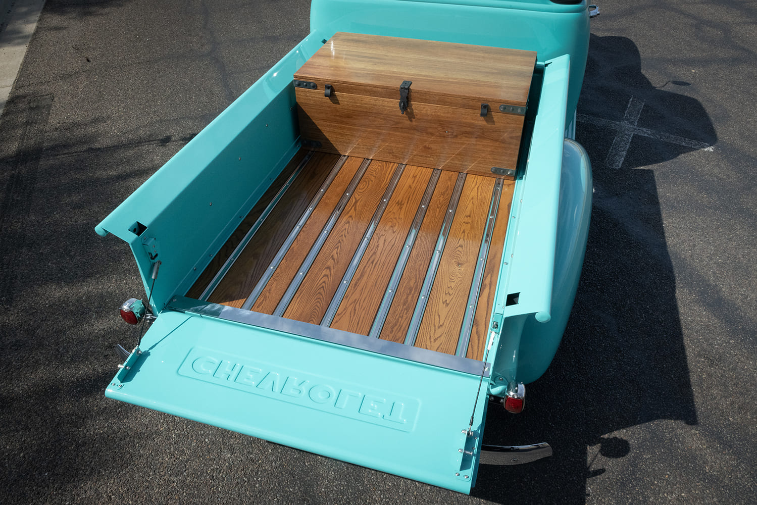 Looking down into the open bed of a turquoise 1953 Chevy pickup showing polished wood planks with stainless strips, a matching wooden chest, and lowered tailgate.