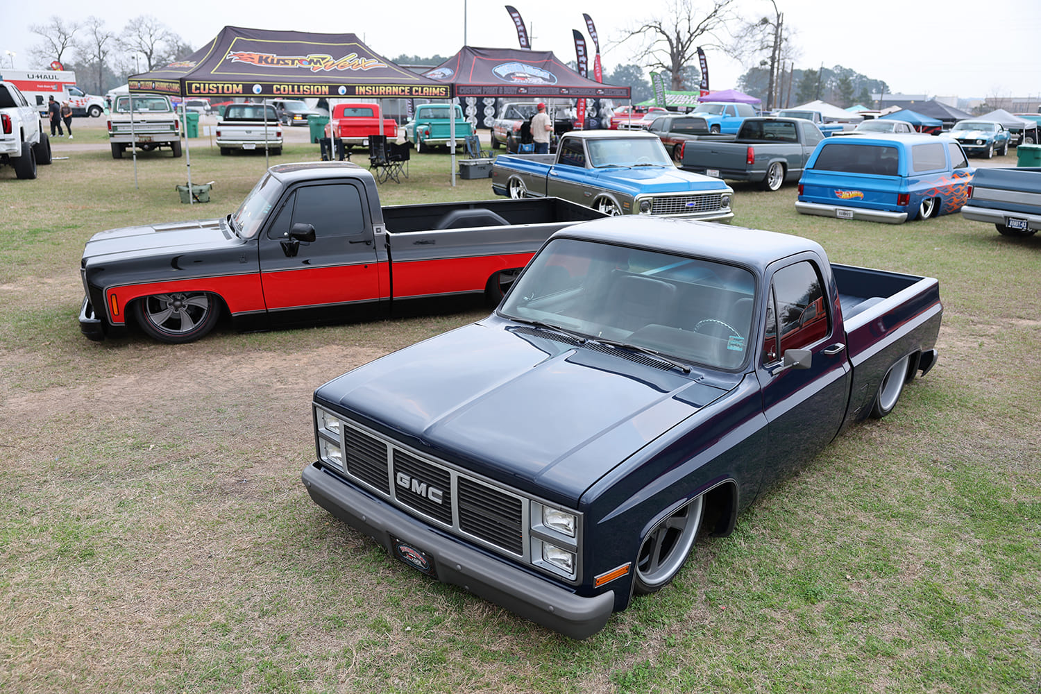 Elevated front-end view of a lowered dark blue GMC squarebody pickup parked next to a two-tone black and red custom truck on the event showfield.