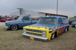 The 2026 Lone Star Throwdown Front three-quarter angle of a customized blue squarebody Chevrolet Suburban featuring airbrushed yellow and orange flames laid out on the grass.