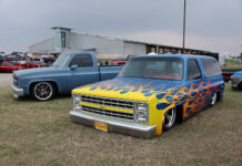 Front three-quarter angle of a customized blue squarebody Chevrolet Suburban featuring airbrushed yellow and orange flames laid out on the grass.