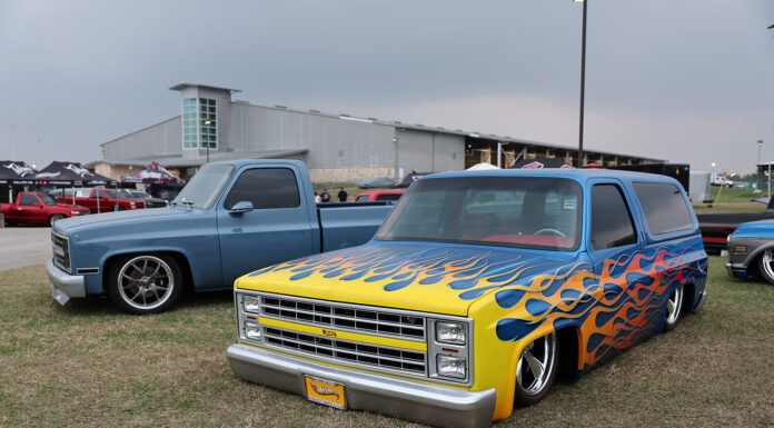 Front three-quarter angle of a customized blue squarebody Chevrolet Suburban featuring airbrushed yellow and orange flames laid out on the grass.