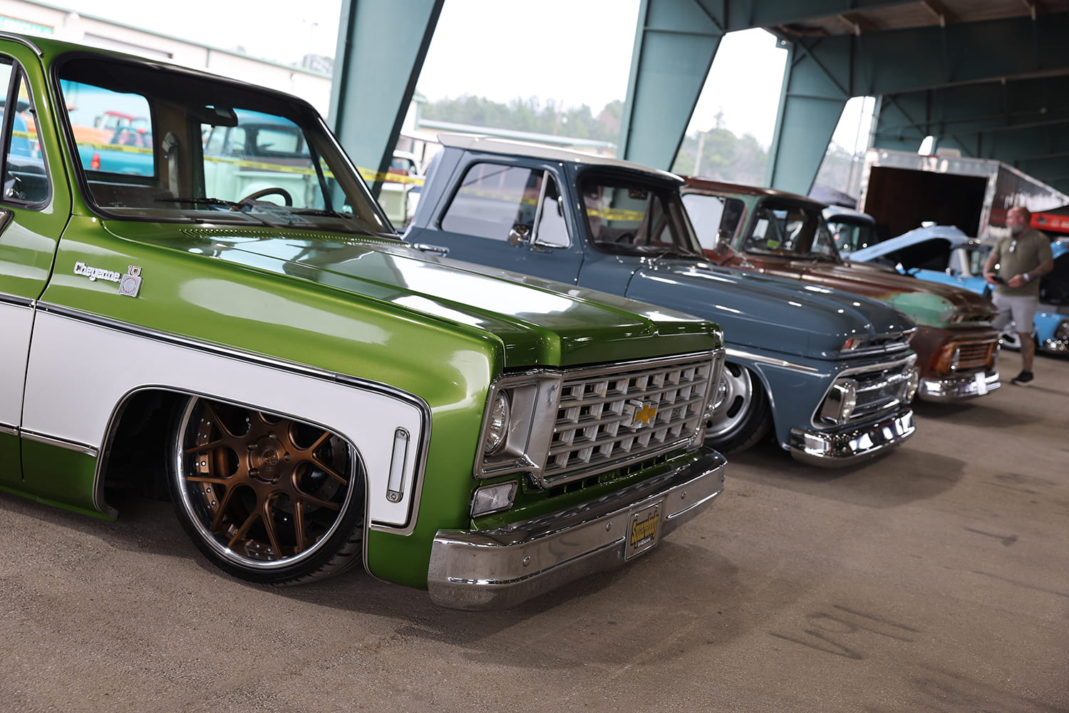 Low-angle front three-quarter view of a lowered lime green and white Chevrolet C10 Cheyenne parked in a row of classic trucks under an arena awning