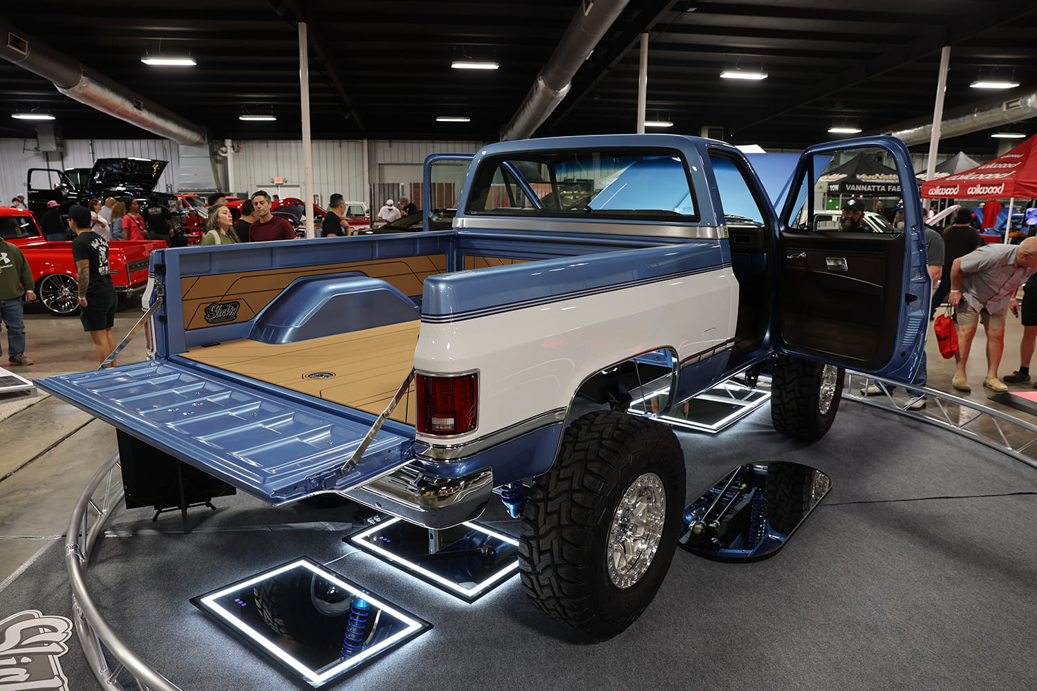 Rear three-quarter view of a lifted blue and white squarebody Chevy pickup with open tailgate, custom finished bed, and polished chrome wheels over floor mirrors.