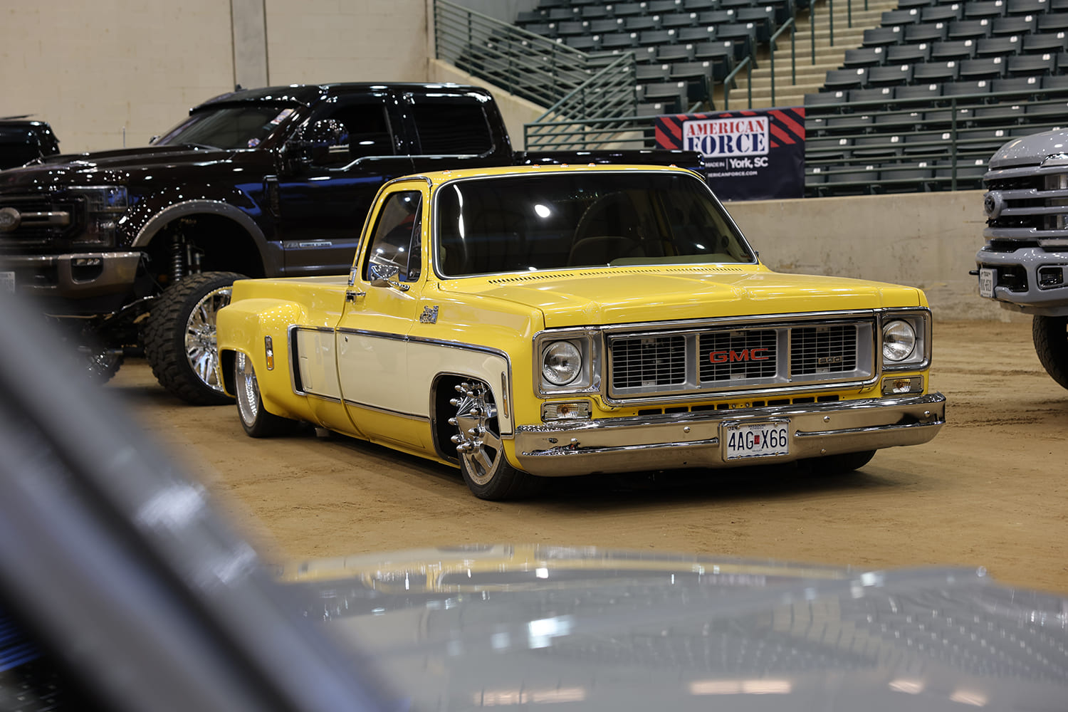 Front three-quarter view of a lowered two-tone yellow and white GMC squarebody dually pickup with spiked lug nuts parked on the arena dirt floor.