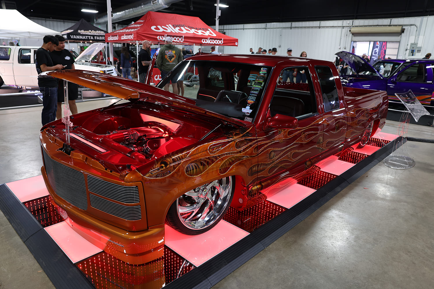 High front three-quarter view of a custom lowered root-beer brown Chevy OBS dually truck with airbrushed flame paint displayed on an illuminated mirror platform indoors
