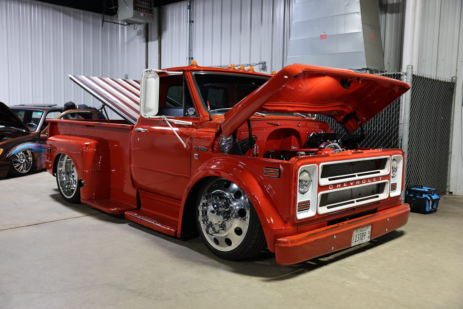 Front three-quarter angle of a custom slammed red Chevrolet C50 cab truck with open hood, polished semi-style wheels, and custom bed cover inside an event arena.