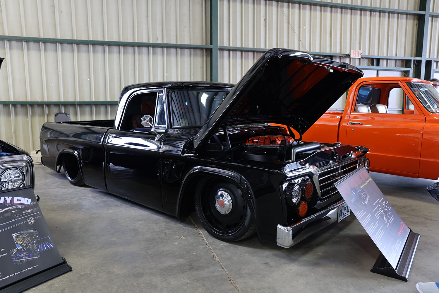 Front three-quarter view of a slammed gloss black vintage Dodge D100 Sweptline pickup with an open hood displaying a modern engine at an indoor show.