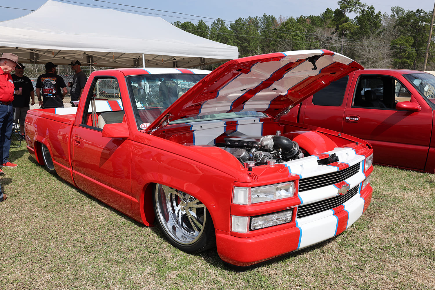 Front three-quarter angle of a lowered red Chevy C1500 OBS pickup with white center racing stripes, open cowl hood, and deep polished billet wheels on grass.