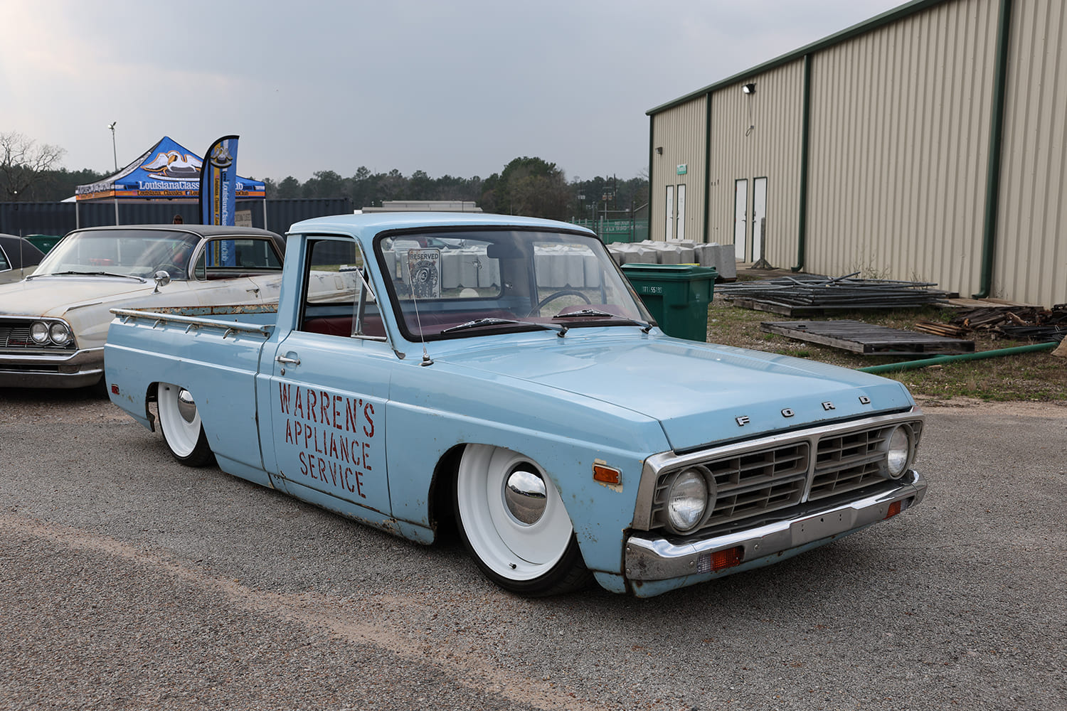 Side three-quarter low profile of a bagged baby blue Ford Courier pickup with vintage door lettering and smoothed white steel-style wheels laid out on asphalt
