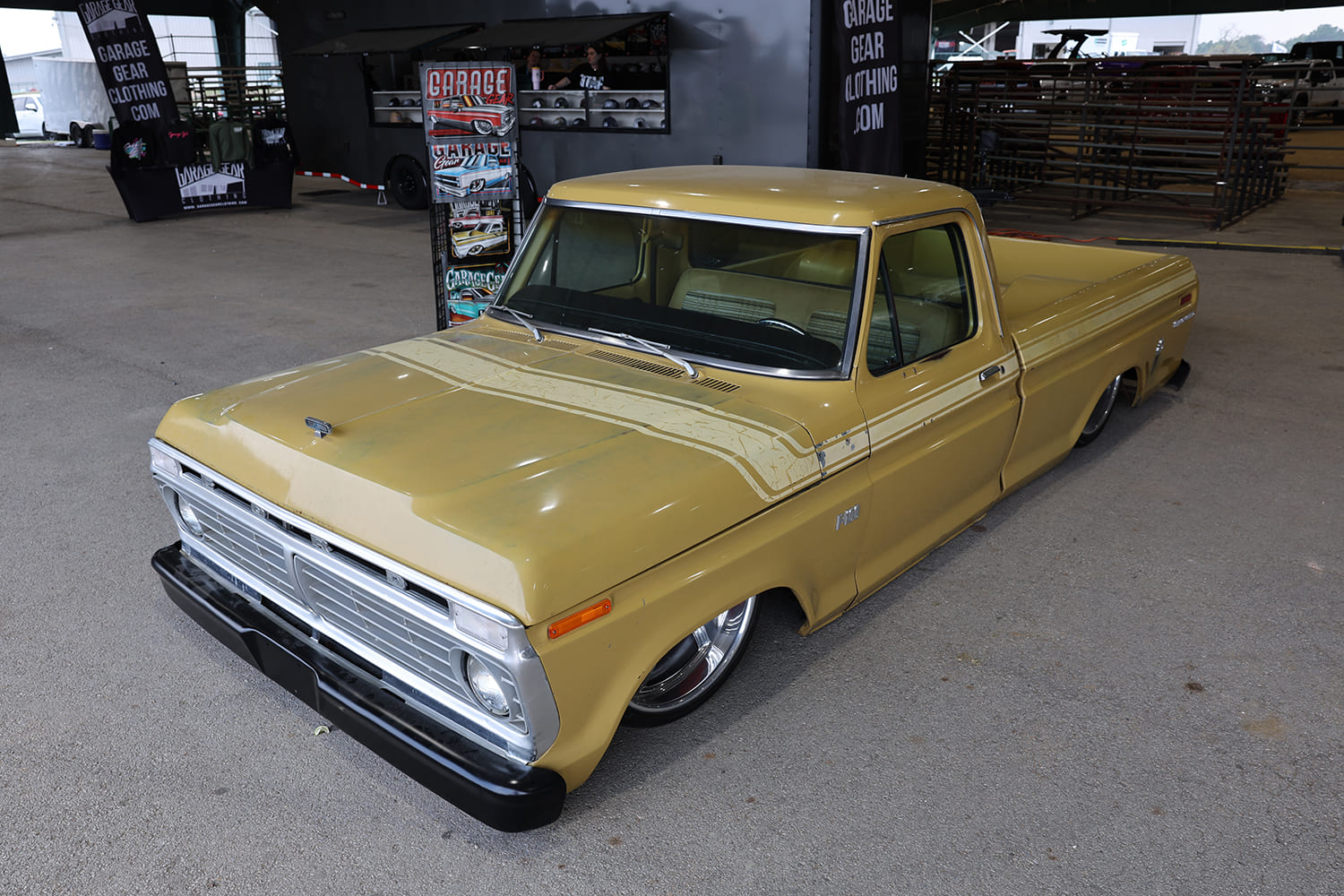 High front three-quarter overhead angle of a lowered vintage yellow-tan Ford F-100 pickup truck with classic body side striping laid out on the arena floor.