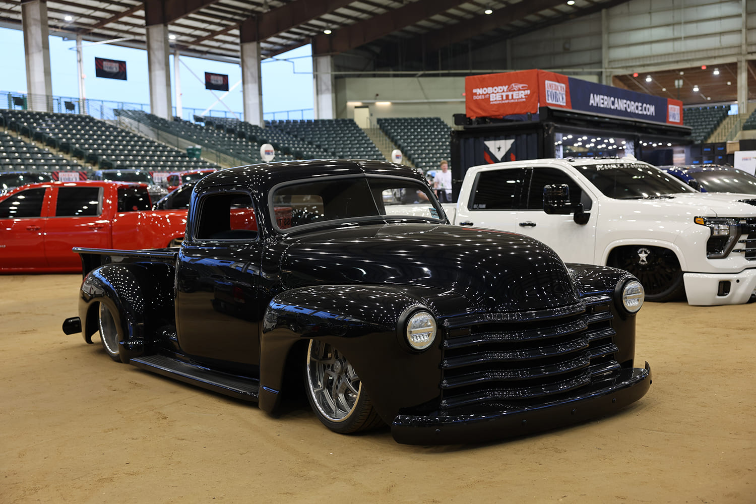 Front three-quarter low angle of a chopped and bagged gloss black vintage Chevy 3100 pickup truck resting on an indoor arena show floor.