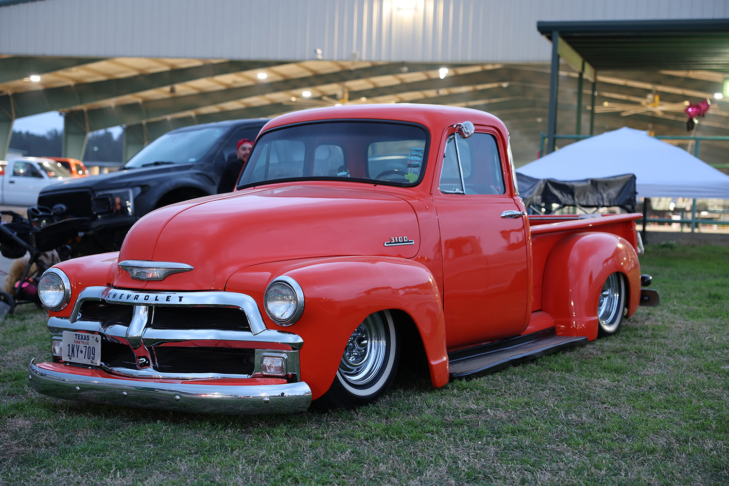Low front three-quarter profile of a bright red custom 1954 Chevy 3100 pickup truck with chrome grille and whitewall tires laid out on grass.