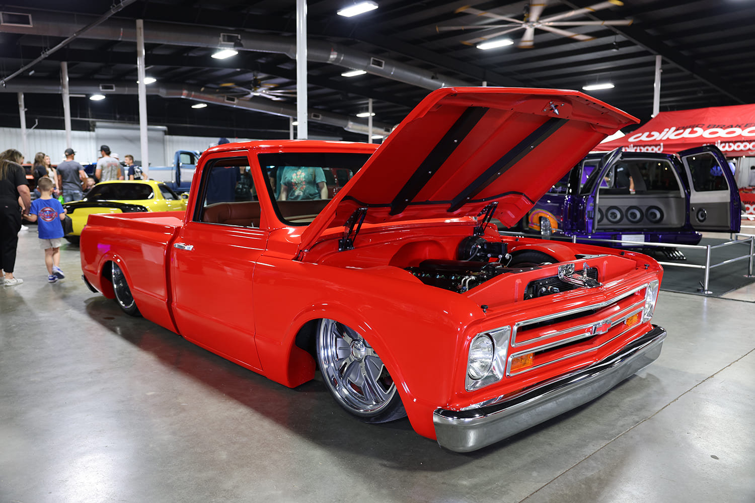 Front three-quarter perspective of a slammed bright red Chevy C10 pickup with open hood revealing a modern V8 and custom inner wheel tubs indoors.