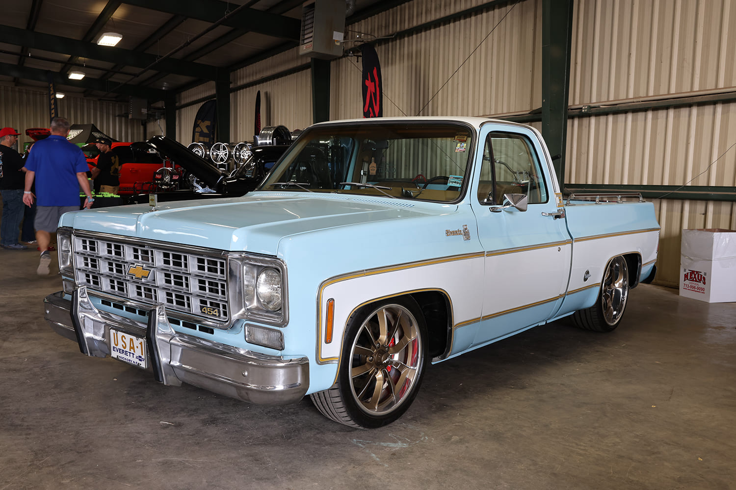 Front three-quarter view of a clean light blue and white two-tone squarebody Chevy C10 pickup with modern bronze wheels parked inside an arena.