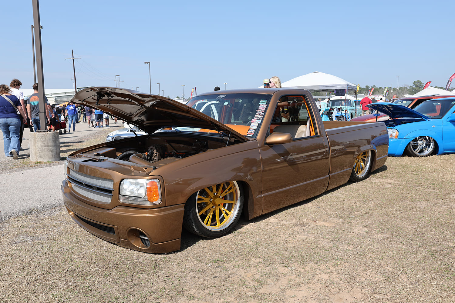 Low-angle front three-quarter shot of a metallic bronze custom Chevy S-10 mini truck with open hood and gold-accented wheels at an outdoor truck show
