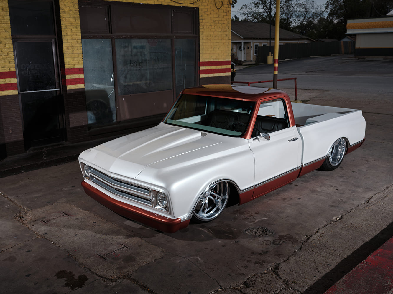 High front three-quarter view of a bagged 1968 Chevrolet C10 pickup finished in pearl white with copper roof and rocker accents parked outside a vintage brick service station.