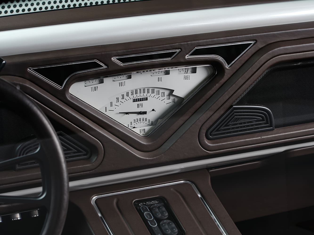Close-up of a custom white-face triangular gauge cluster set into a molded brown leather dashboard with an integrated AccuAir e-Level air suspension keypad.