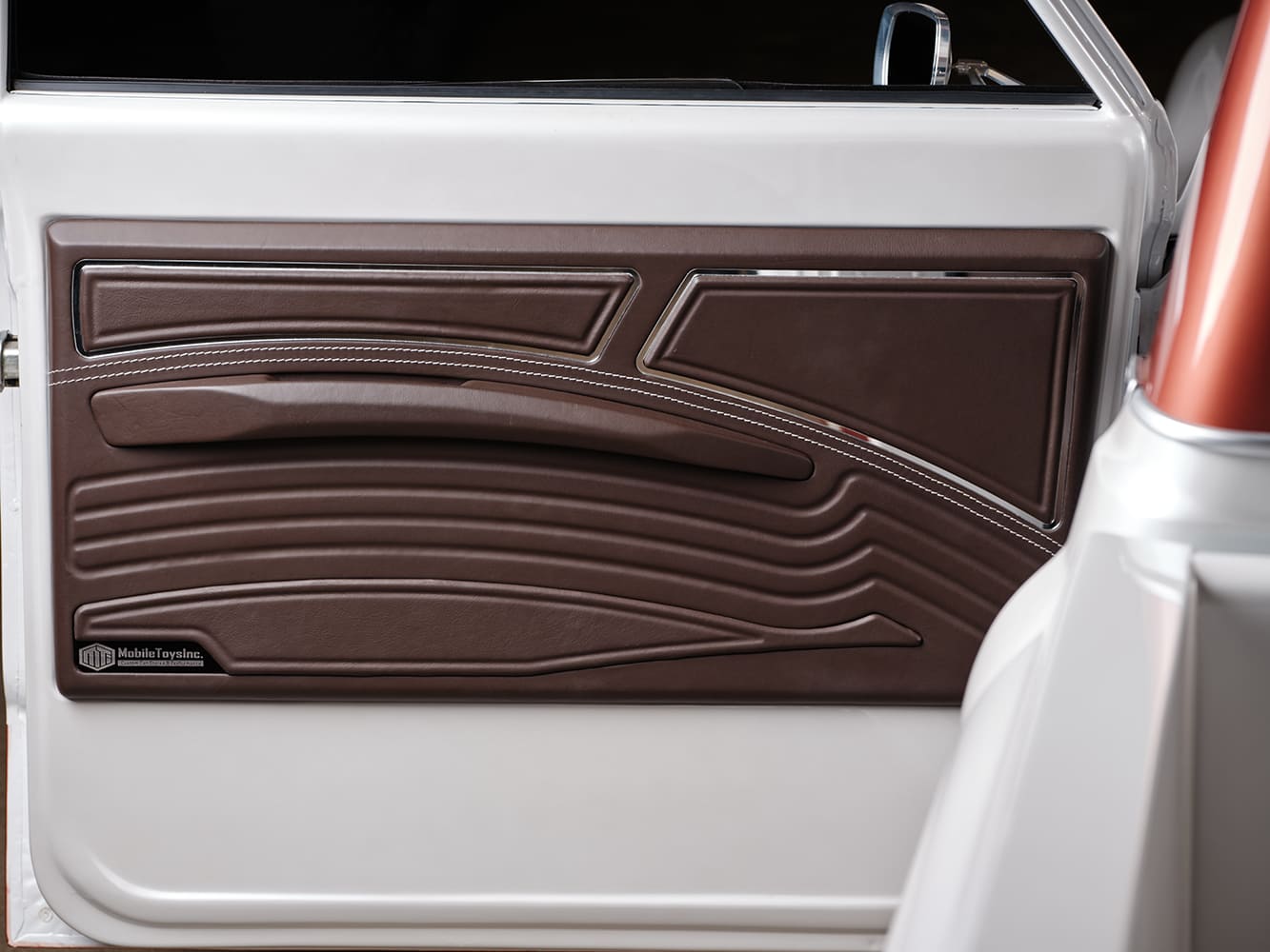 Close-up of the sculpted two-tone brown leather door panel with Mobile Toys Inc badge, contrast stitching, and integrated armrest.