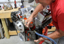 Mechanic using an open-end wrench to adjust mounting bracket tension on a Saginaw P-pump mounted to an orange small-block Chevy V8 on an engine stand.