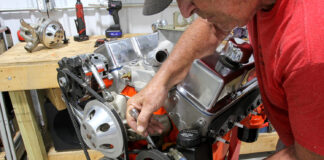 Mechanic using an open-end wrench to adjust mounting bracket tension on a Saginaw P-pump mounted to an orange small-block Chevy V8 on an engine stand.