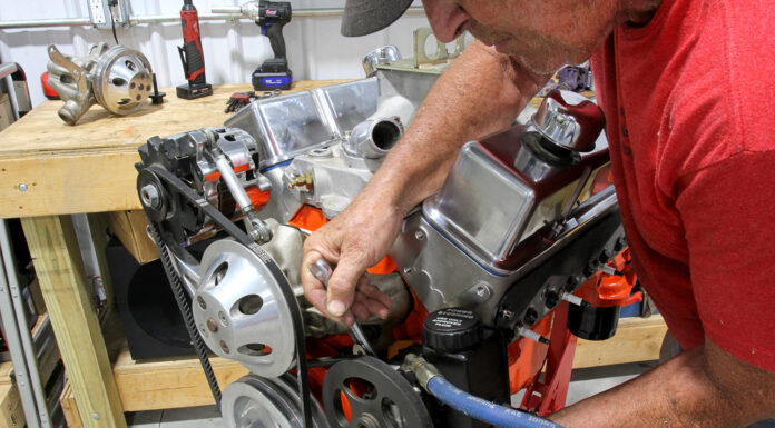 Mechanic using an open-end wrench to adjust mounting bracket tension on a Saginaw P-pump mounted to an orange small-block Chevy V8 on an engine stand.