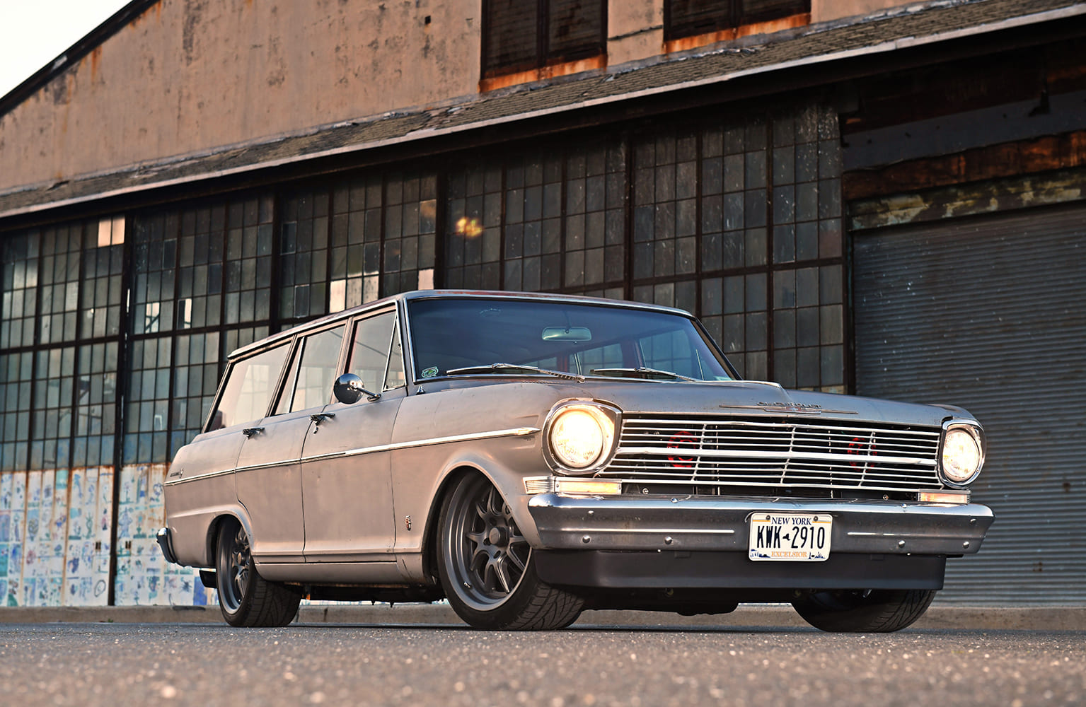 Low-angle front three-quarter view of a patina silver 1962 Chevrolet Nova station wagon restomod with glowing headlights parked outside an industrial warehouse building.