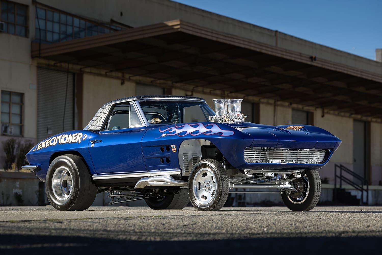 Low-angle front three-quarter view of a blue 1964 Chevy Corvette gasser with custom flames, straight front axle, and velocity stacks.