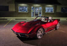 Front three-quarter night view of a custom candy red 1972 Chevrolet Corvette roadster with cowl hood and side exhaust pipes parked outside a brick building.