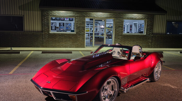 Front three-quarter night view of a custom candy red 1972 Chevrolet Corvette roadster with cowl hood and side exhaust pipes parked outside a brick building.