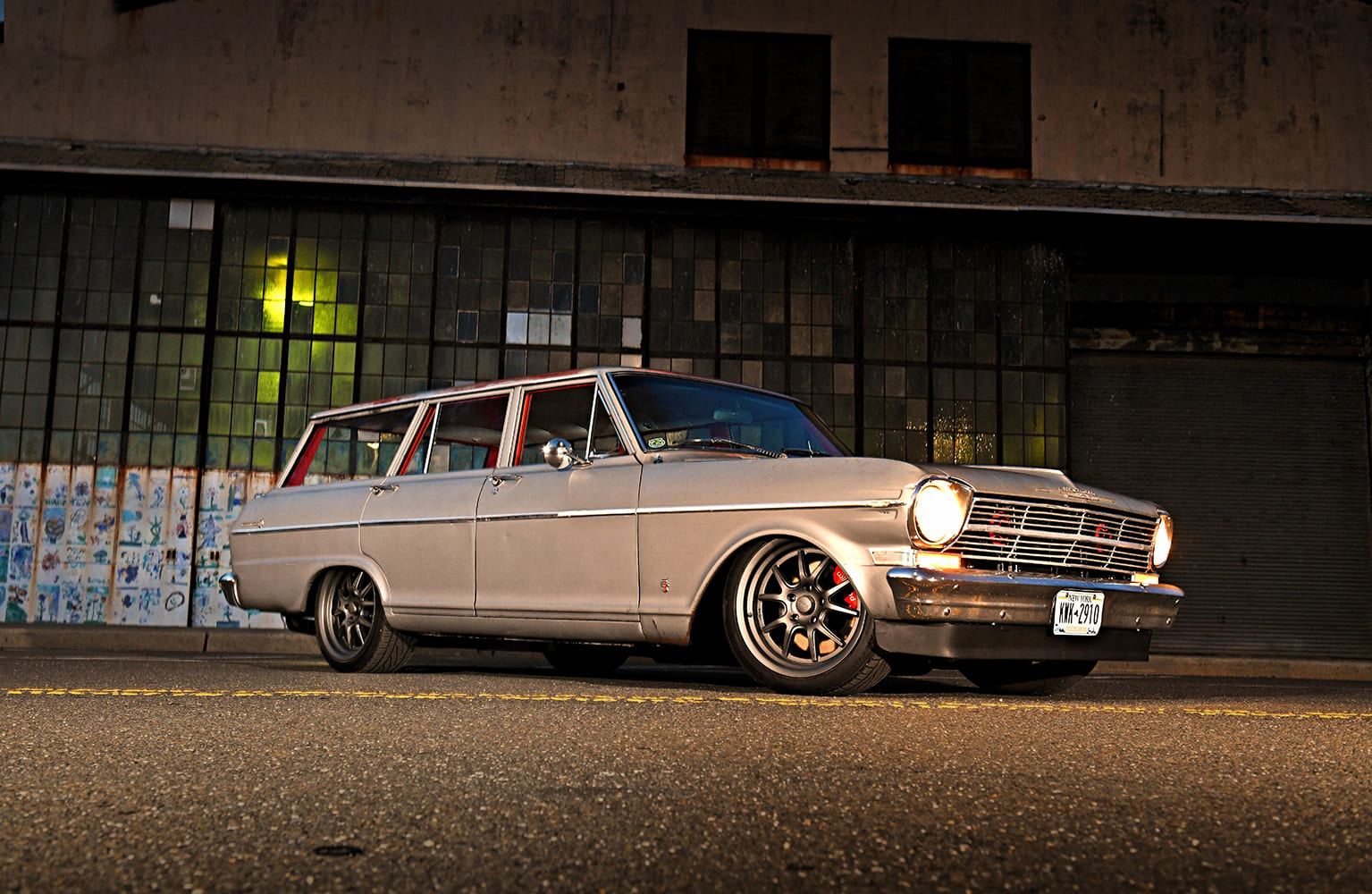 Front three-quarter angle of a lowered silver 1962 Chevy Nova wagon on gunmetal wheels illuminated by warm streetlamps in front of a weathered brick and glass factory.