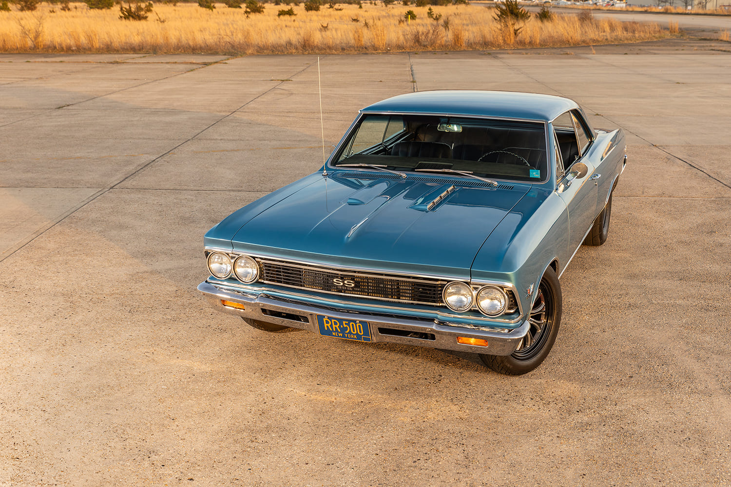 Elevated front three-quarter perspective looking down across the hood louvers, windshield, and blue metallic paint of the 1966 Chevelle parked on an airfield ramp.