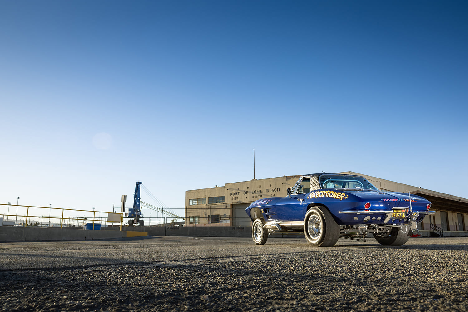 Wide industrial shot of a blue 1964 Chevy Corvette gasser parked outside a warehouse at the Port of Long Beach.