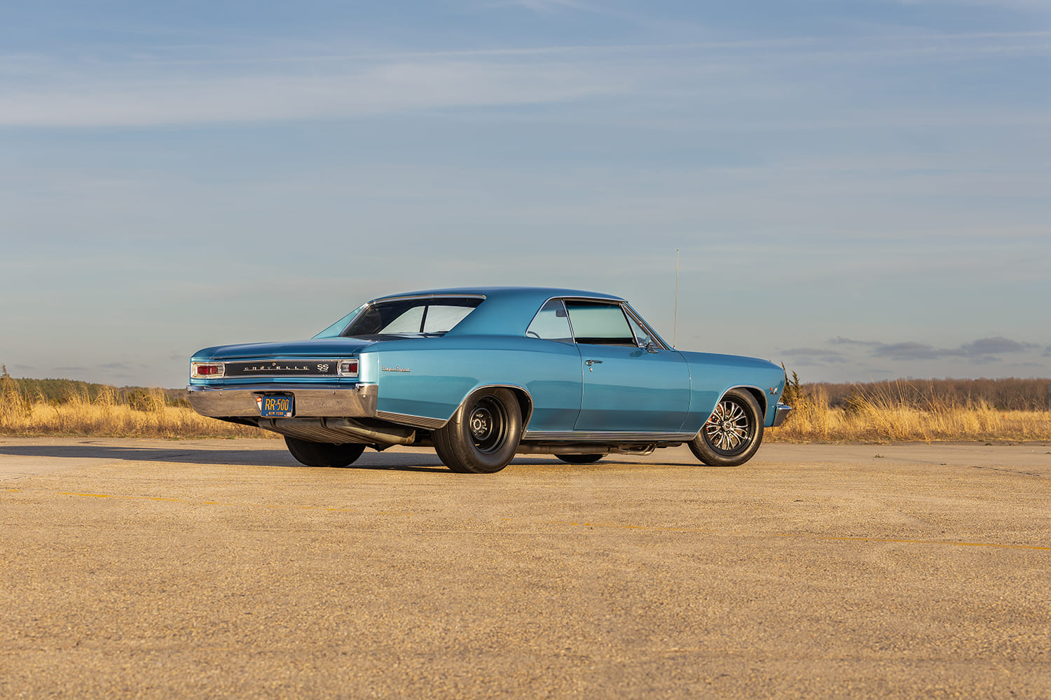 Rear three-quarter view showing the wide rear drag radial tires tucked into the wheelwells, chrome rear bumper, and horizontal taillights of the blue 1966 Chevelle.