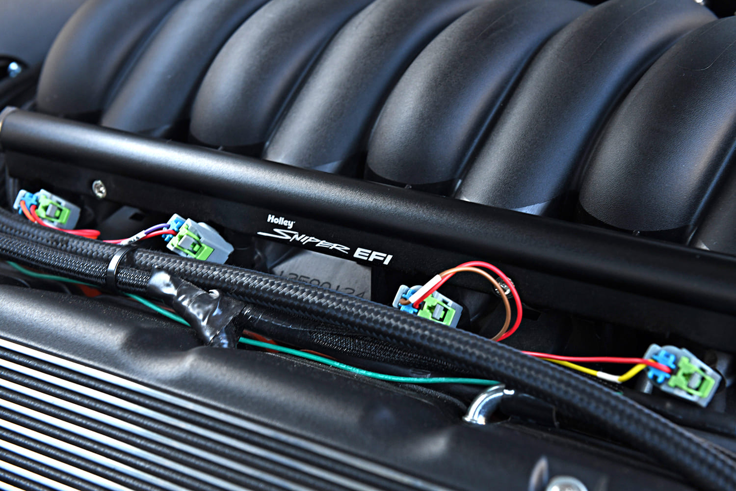 Close-up macro view of the black anodized Holley Sniper EFI fuel rail, braided fuel crossover line, and injector harness on the LS engine intake manifold.