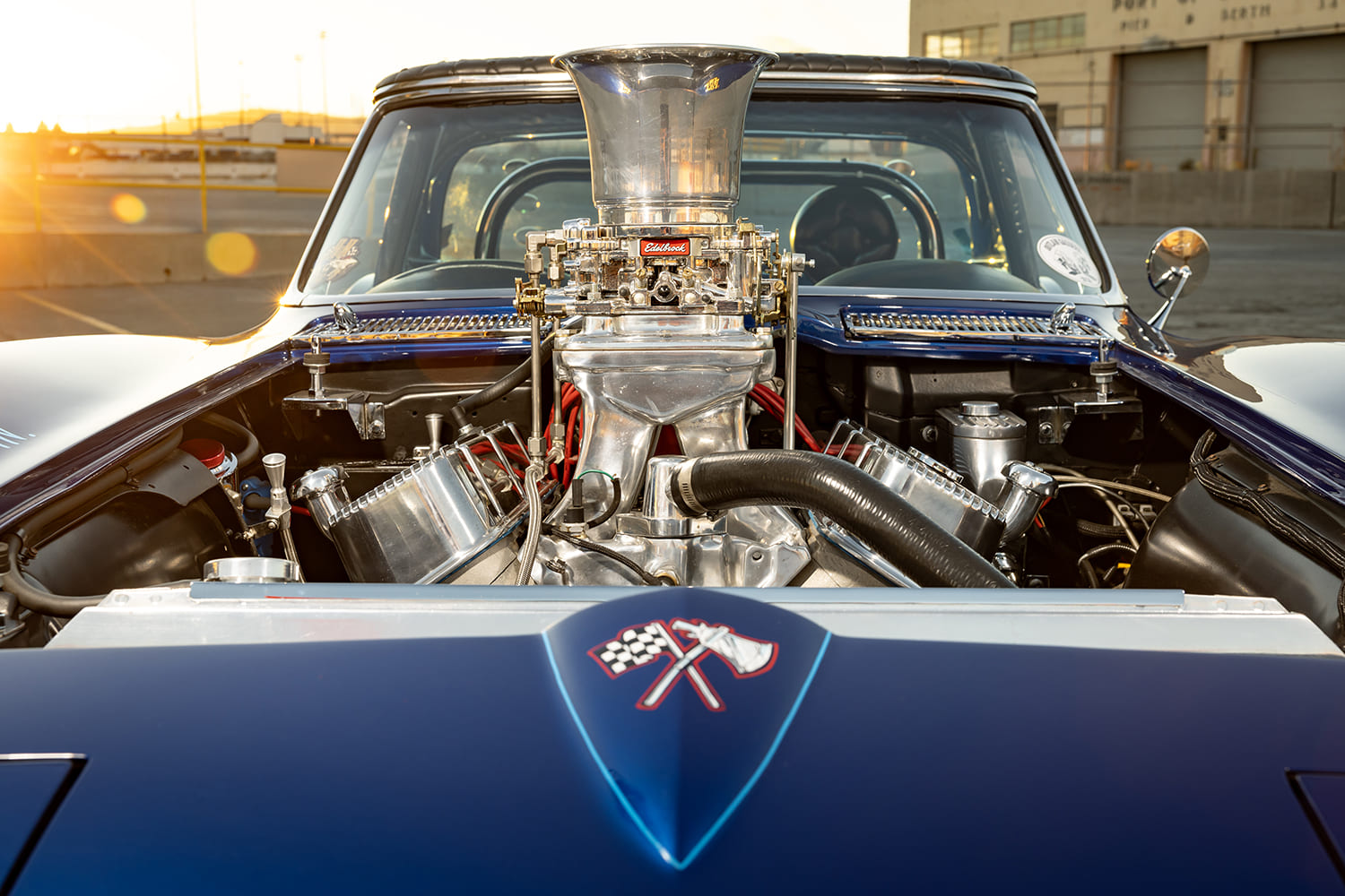 Front overhead perspective of a blue 1964 Corvette gasser nose emblem and towering dual carburetors back-lit by sunset.