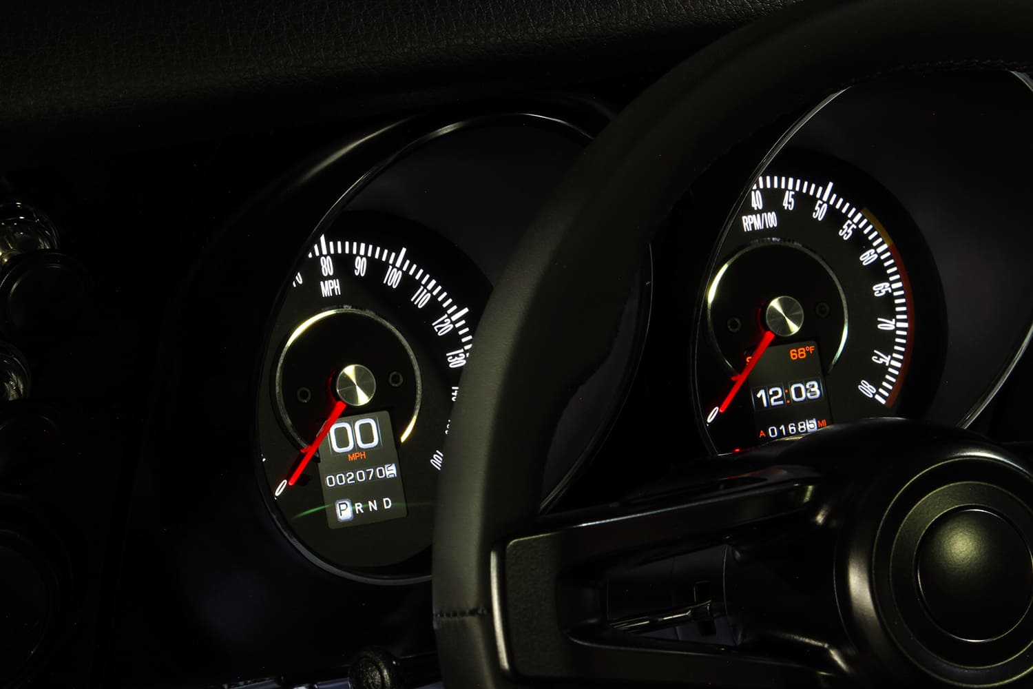Driver viewpoint of retro-styled electronic speedometer and tachometer gauges with digital displays framed behind the steering wheel.